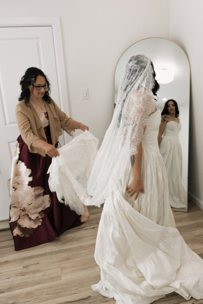 A woman in glasses and a floral dress assists a bride trying on a white lace wedding gown and veil in front of a mirror.