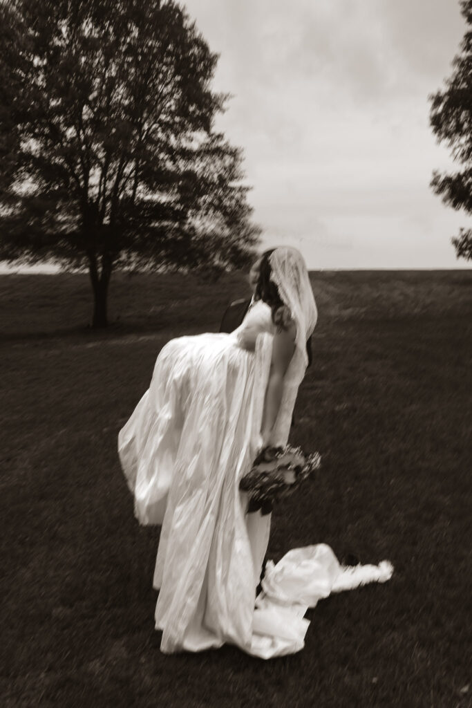 Sepia-toned image of a person in a wedding dress being carried outdoors, with trees in the background.