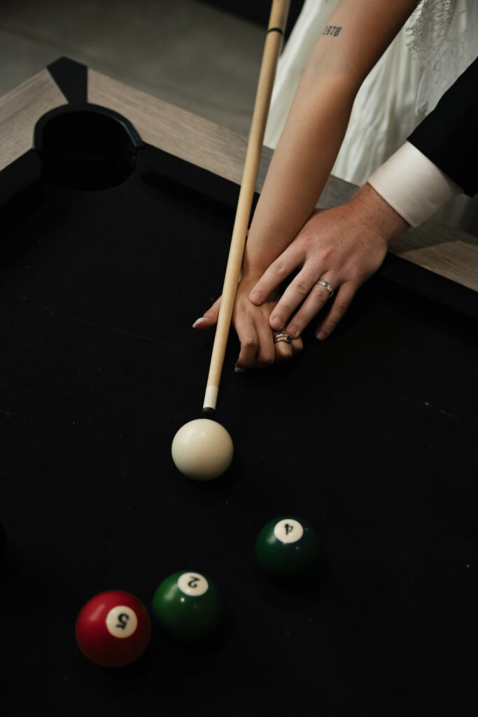 Bride and groom holding hands on pool table with cue and balls.