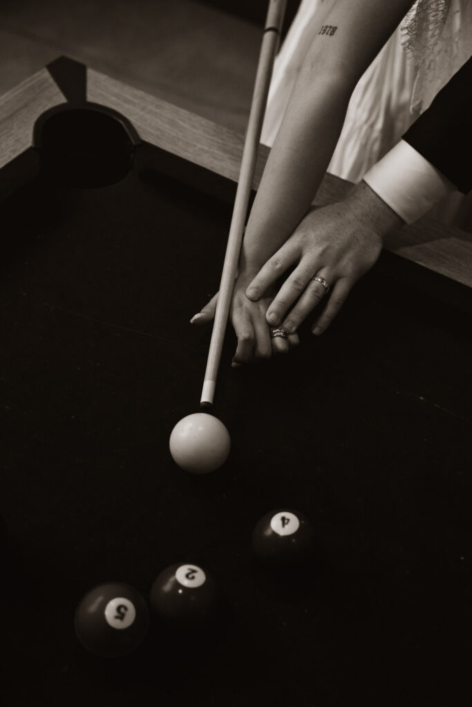 Bride and groom's hands together on a pool table, with a cue stick and balls visible.