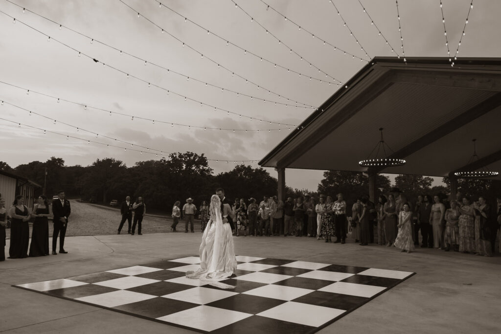 Bride and groom dancing on a checkered floor under string lights with guests watching, outdoor wedding reception.