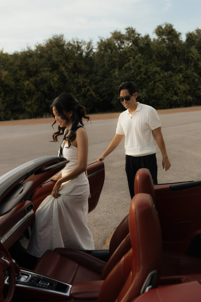Smiling woman in white dress getting into a red convertible sports car while a man in sunglasses holds the door open.