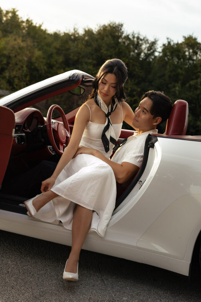 Elegant couple in white attire sitting in a vintage convertible car with lush greenery background.