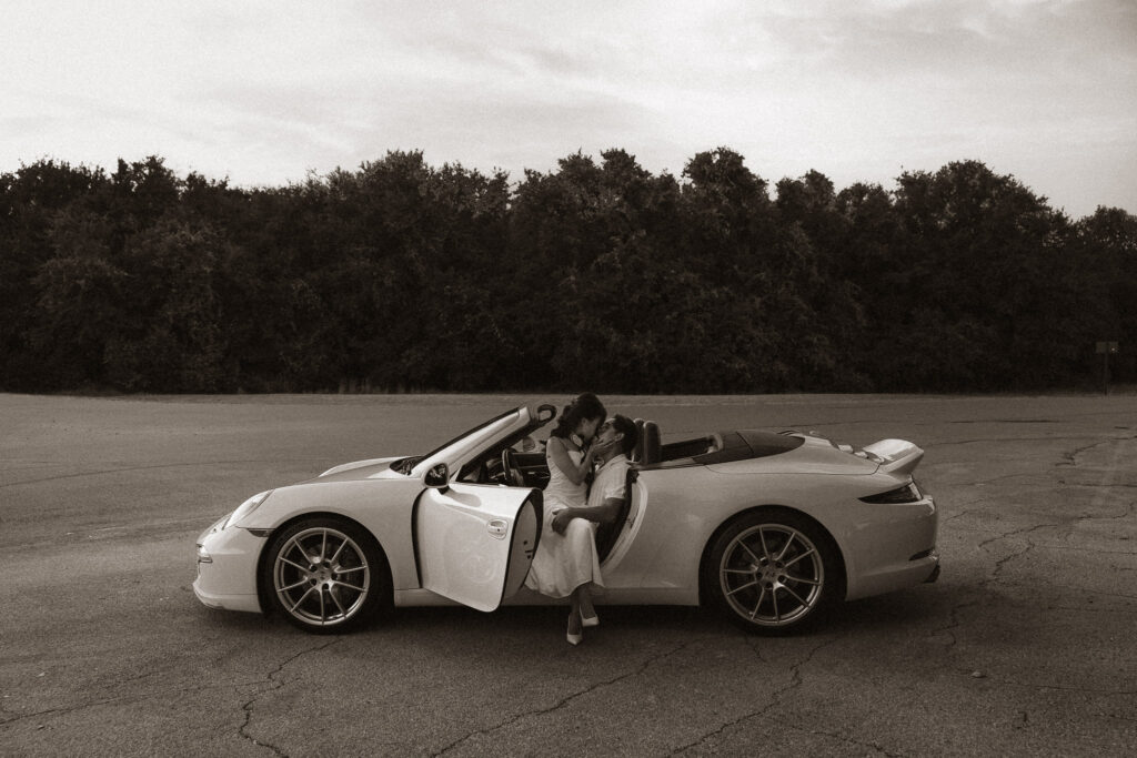 Monochrome image of a couple kissing in a white Porsche convertible with the door open, parked on an asphalt road with trees in the background.