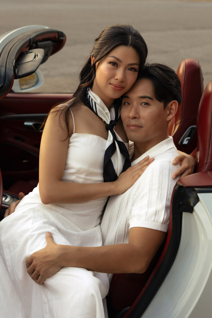 An elegant couple in a luxury convertible. The woman, in a white dress and silk scarf, embraces the man, who wears a white polo.