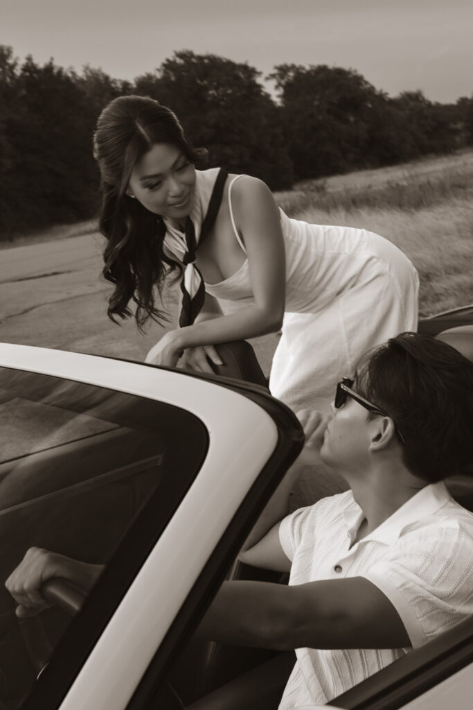 A woman in a white dress leans over to speak to a man in sunglasses driving a convertible, sepia tone.