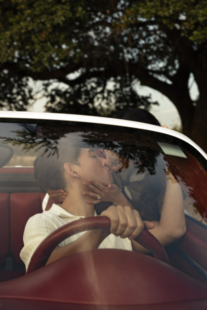 Engaged couple kissing in a vintage car. A woman leans over the windshield to kiss her fiance who is behind the steering wheel.