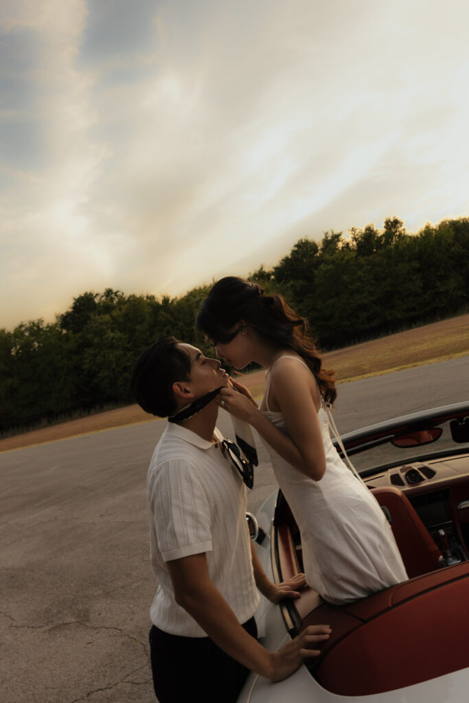 A stylish couple shares an intimate moment, with the woman in a white dress adjusting the man's scarf next to a vintage car.