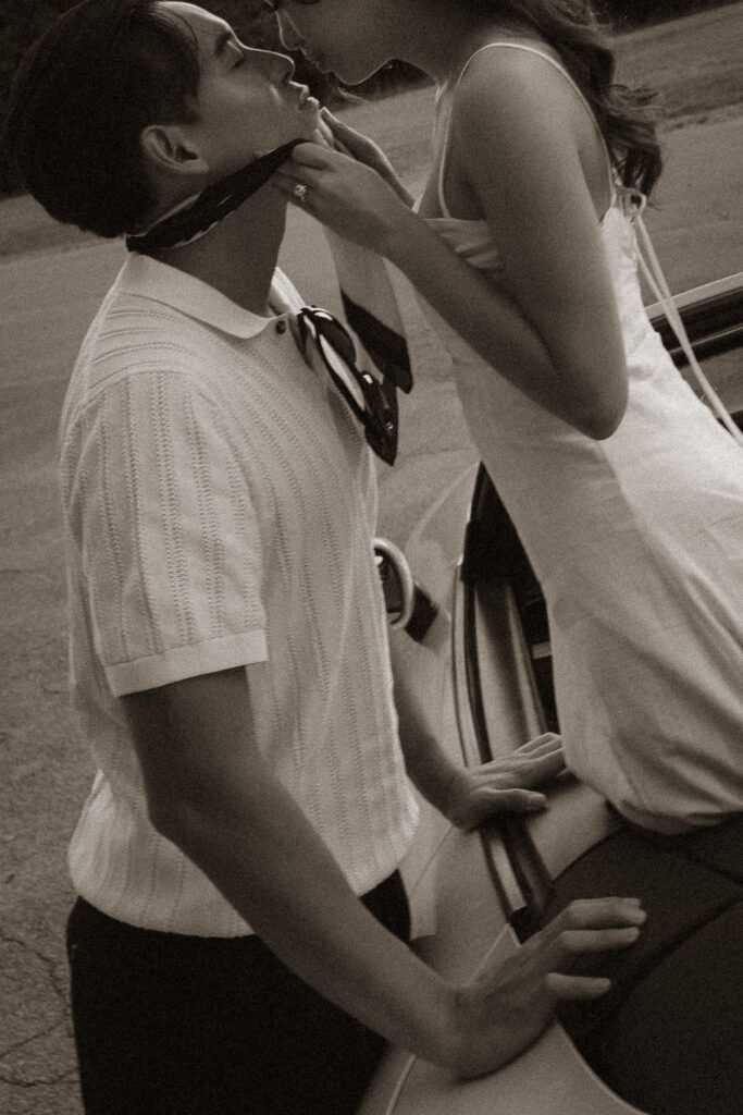 Engaged couple embracing by a vintage car, showing off engagement ring. Man in polo, woman in white dress.