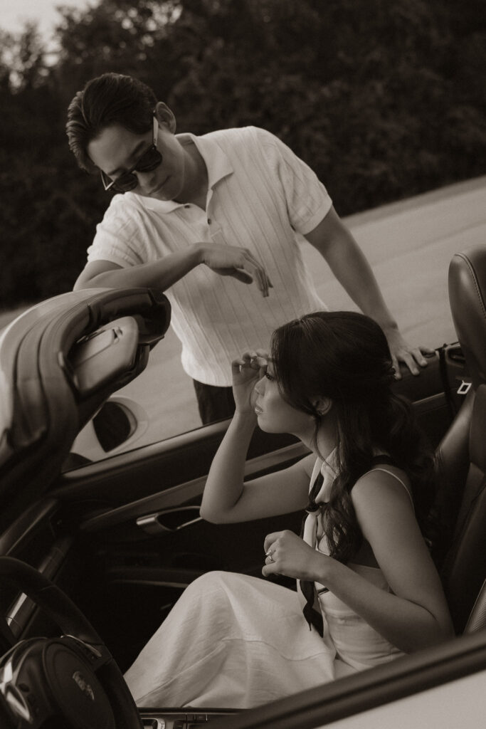 Monochromatic image of a man in sunglasses leaning on a convertible while a woman applies makeup in the passenger seat.