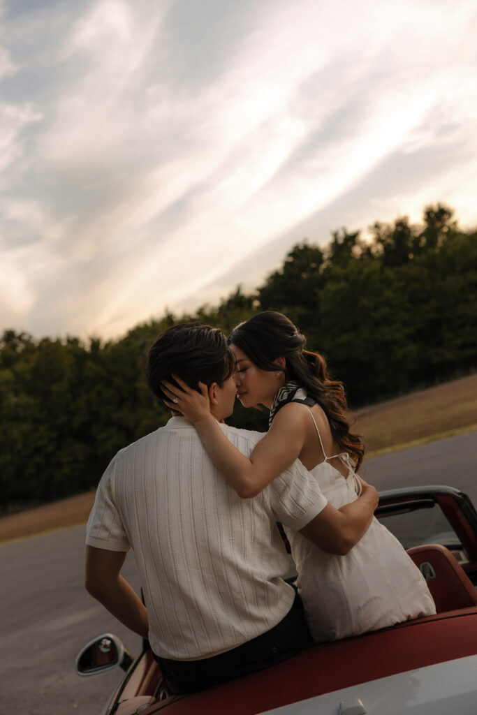 An engaged couple embraces and kisses while sitting on the trunk of a red vintage car, surrounded by lush green trees and a soft sunset sky. The woman wears a white dress and the man a striped polo.