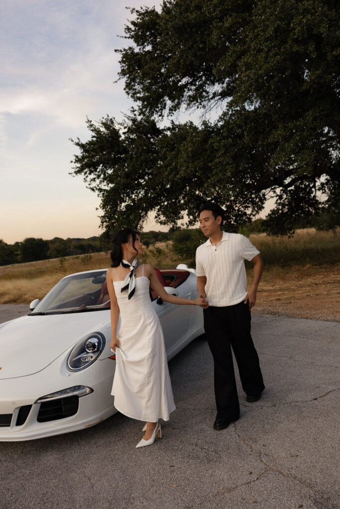 An Asian couple holding hands next to a white Porsche convertible, with the woman in a white dress and the man in a white polo.