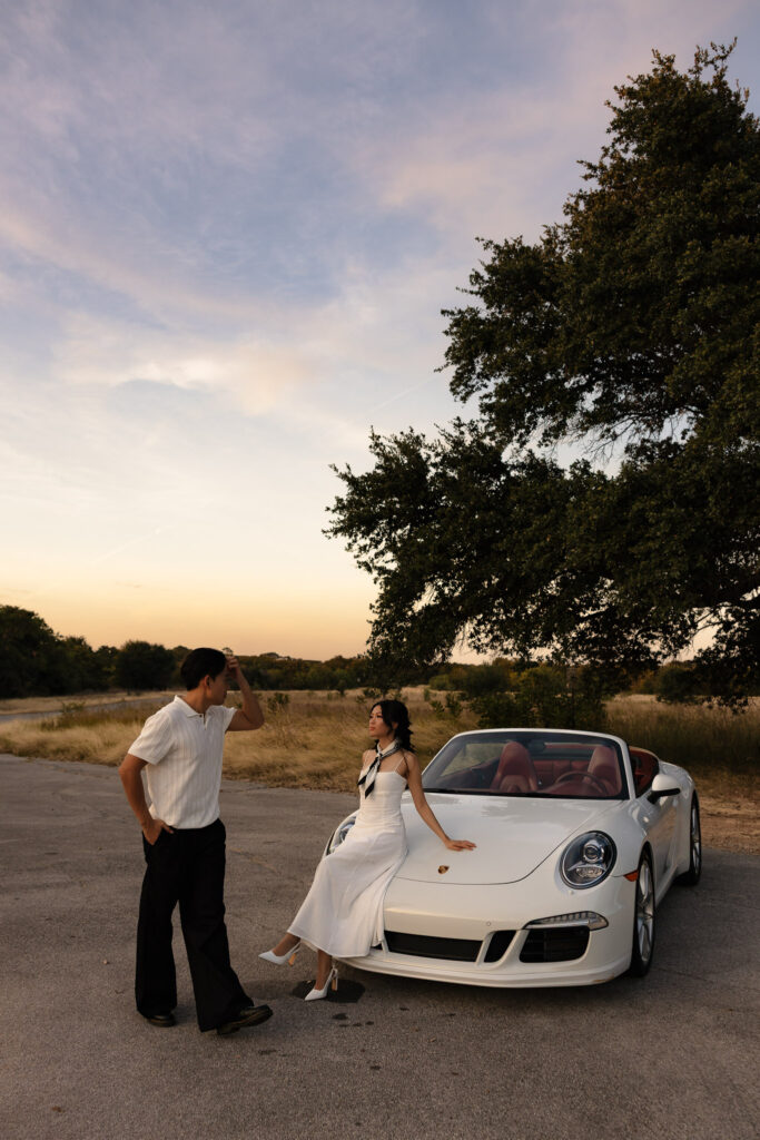 Engaged couple with vintage white Porsche convertible at sunset. Woman in white dress poses on car, man in black pants stands by.