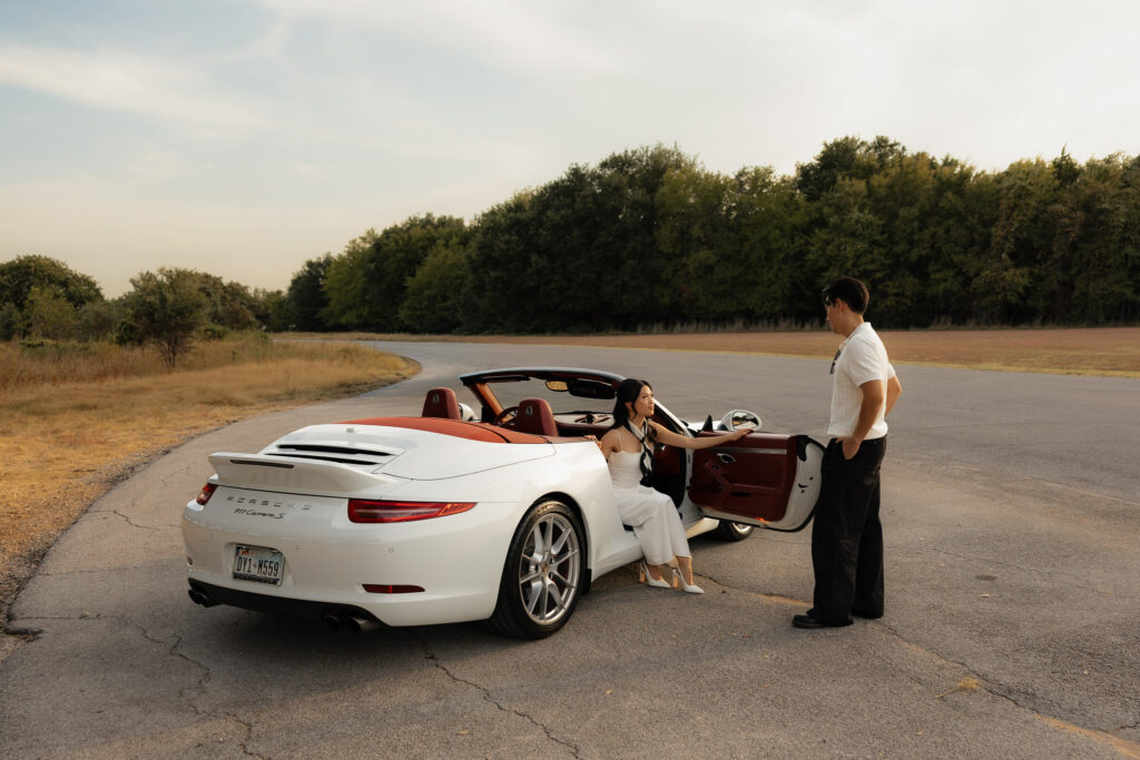 Engaged couple with white Porsche 911 Carrera S convertible, orange interior, on a winding road with trees and sky.