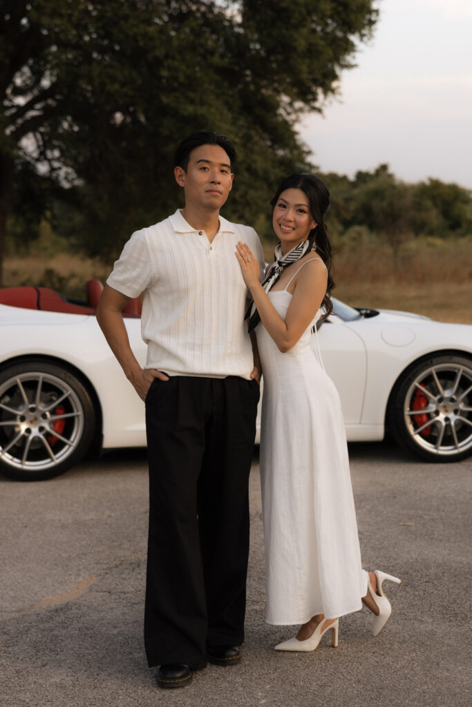 An elegant couple poses next to a white Porsche convertible. The woman wears a white dress and scarf, the man a cream polo and black pants.
