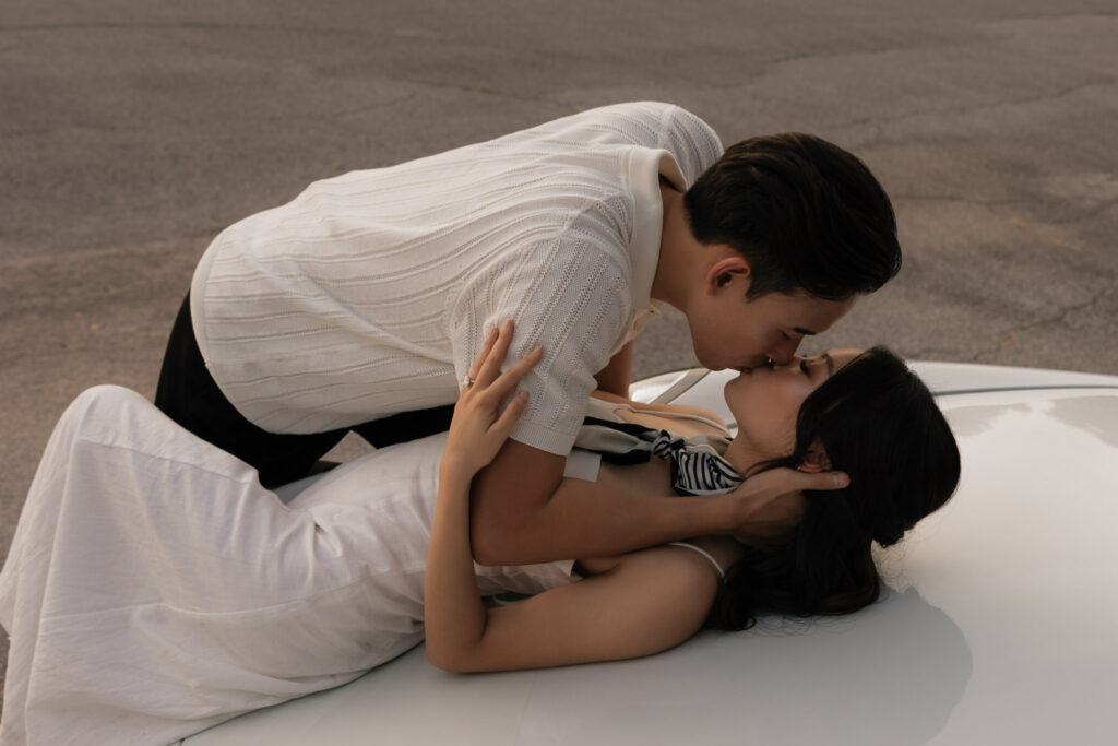 An Asian couple passionately kissing on a white vintage car. The woman in a white dress is lying on the hood, the man in a white polo shirt leans over her.