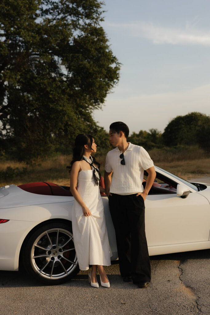 Engaged couple in a vintage white car. The man is wearing a white shirt and black trousers, while the woman is wearing a white dress and black shoes. They are looking at each other, in love, leaning on their vintage car. The background is a forest.