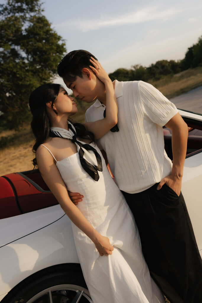 A romantic couple in white outfits leaning against a classic white car, gazing into each other's eyes.