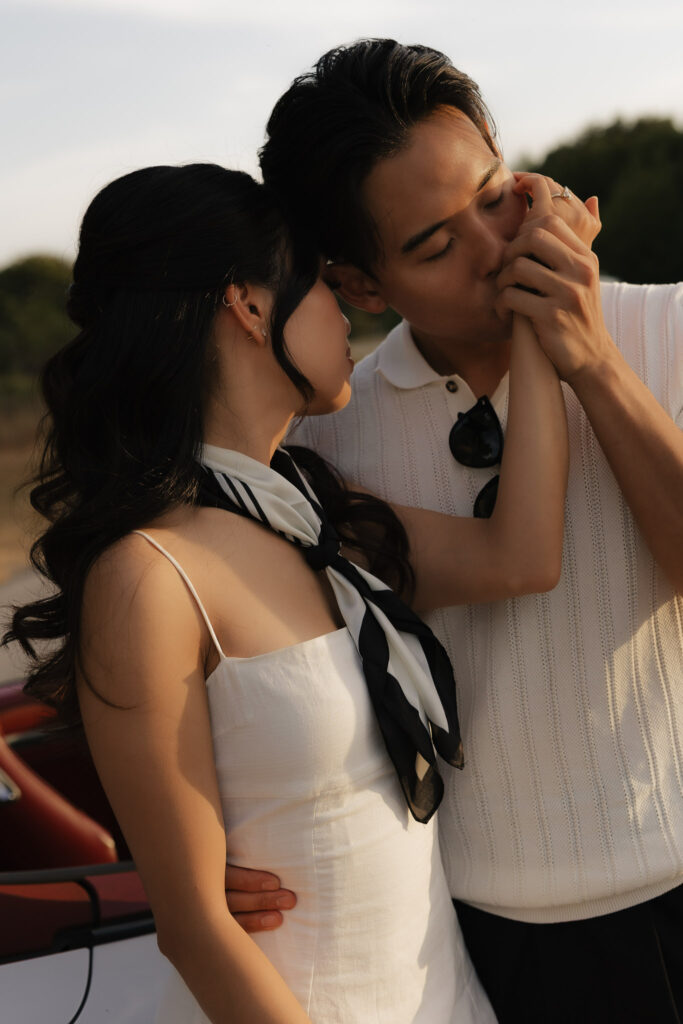 An affectionate couple in elegant attire, with the man tenderly kissing the woman's hand, against a soft, golden hour backdrop.