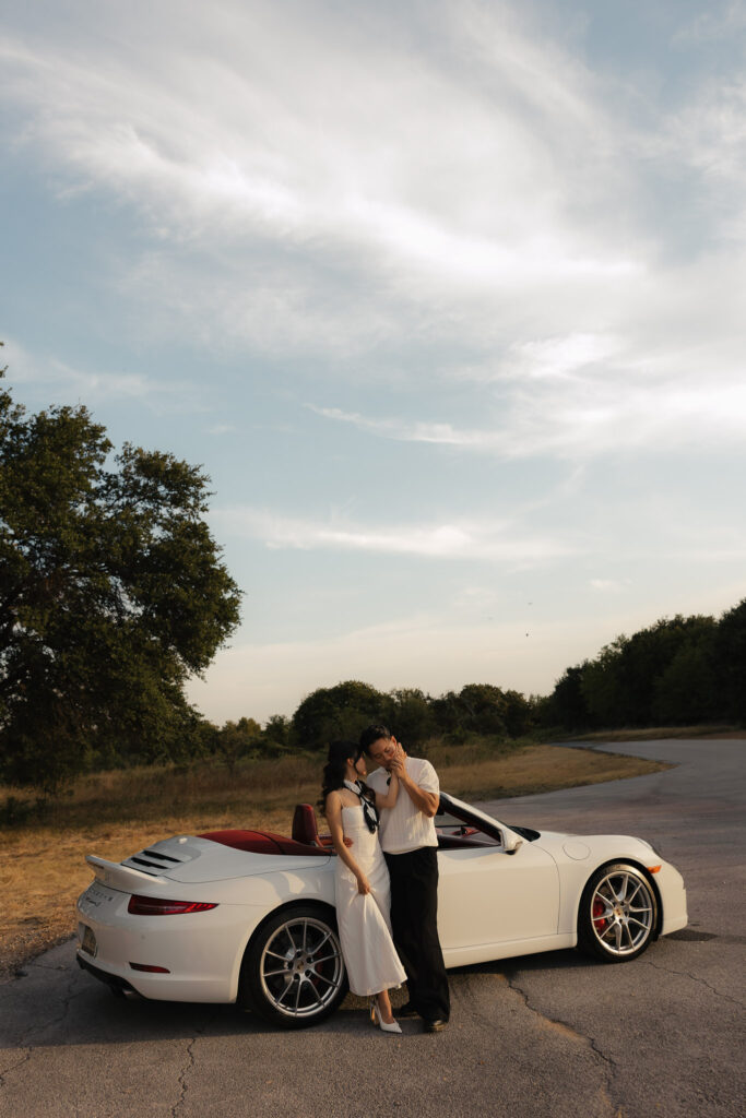 Engaged couple in elegant attire pose intimately beside a white Porsche convertible on a scenic country road at sunset.