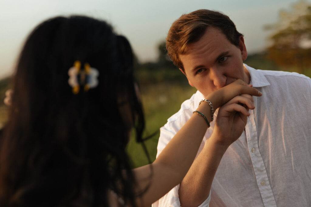 Man gently kissing woman's hand during lakeside engagement at sunrise.