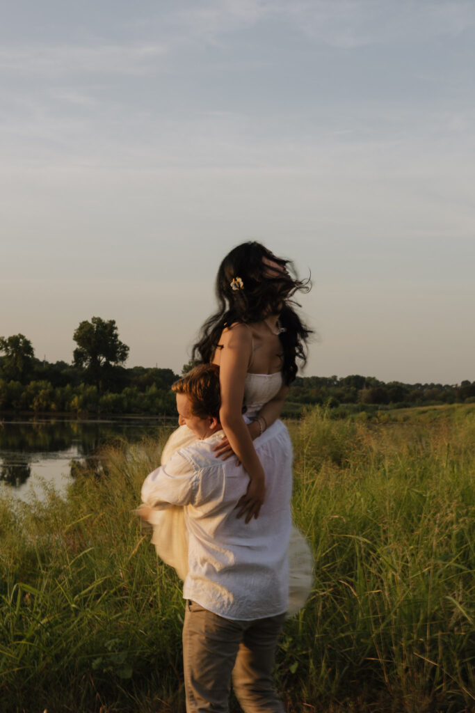 A couple enjoying a romantic moment by a lakeside at sunrise. The woman is playfully lifted by her partner, surrounded by lush greenery.